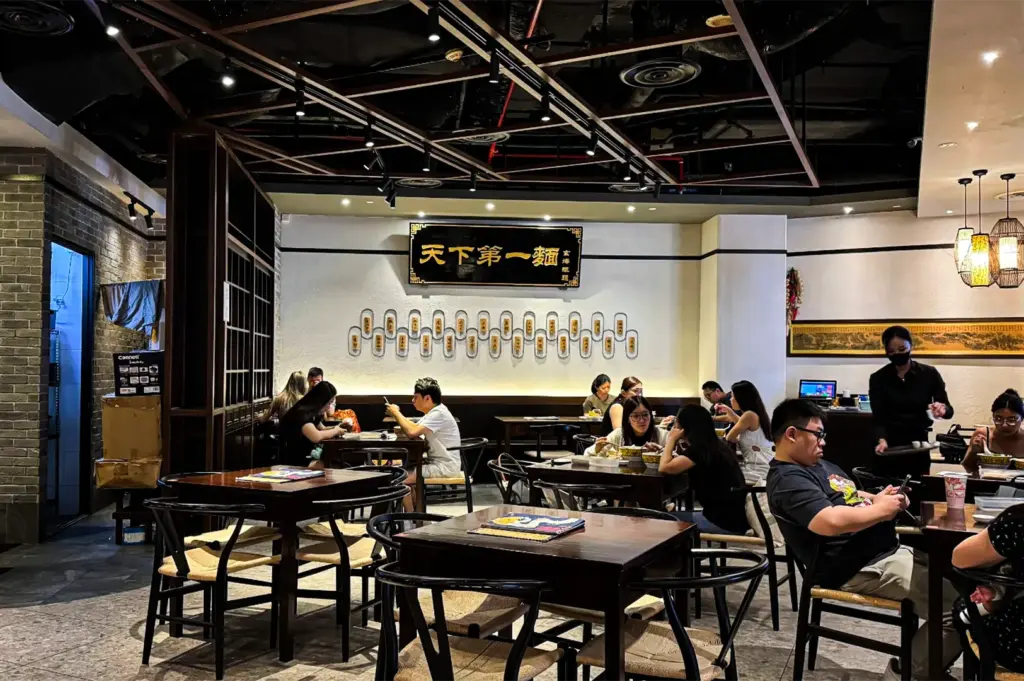 An interior view of a bustling, brightly lit casual Chinese restaurant filled with patrons dining at dark square wooden tables paired with woven-seat chairs. The white back wall features a prominent horizontal black sign with gold Chinese characters reading "天下第一麵," above a decorative row of small vertical wooden menu plaques. Overhead, an open industrial ceiling is crisscrossed by dark wooden beams and tracking lights, while a standing waitress in a black uniform attends to a table on the right near hanging decorative lantern lights.