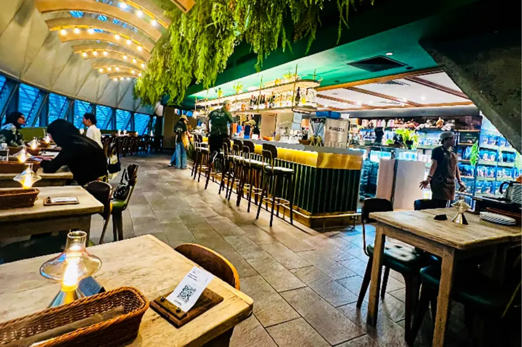 An interior view of a modern, stylish restaurant featuring a long, illuminated bar counter with green paneling and gold trim, lined with high barstools. Lush green foliage hangs densely from a section of the ceiling over the bar area, contrasting with a curved, light-colored architectural ceiling illuminated by rows of small lights on the left. Patrons sit at rustic wooden tables equipped with small, glowing glass lamps and woven baskets, while staff move about the tiled floor near a glass display case in the background.