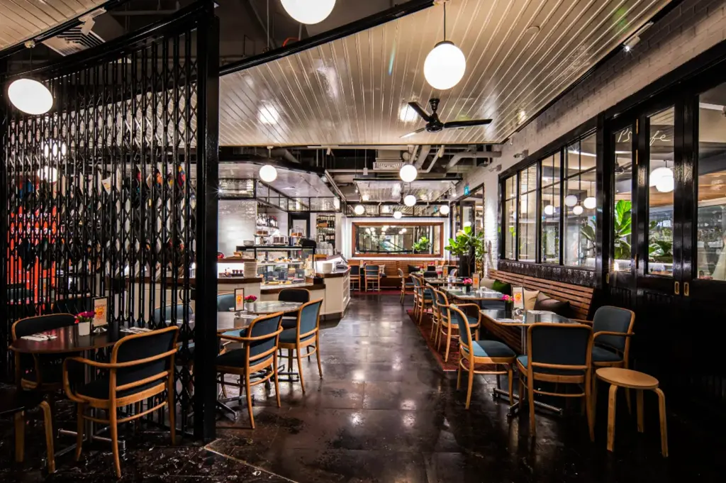 An interior view of a spacious, modern cafe with polished dark flooring and a glossy white-slatted ceiling dotted with glowing globe pendant lights. On the left, a black accordion-style metal gate partially screens off a dining area featuring round tables and blue-cushioned wooden chairs, while a pastry display counter stands further back. The right side features a long wooden banquette lined with small tables, matching blue chairs, and expansive black-framed windows that look out into another brightly lit interior space.