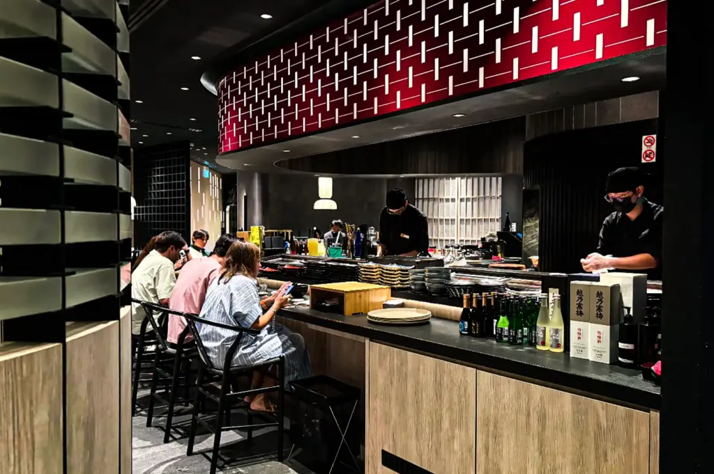 An interior view of a modern sushi or conveyor belt restaurant featuring patrons seated along a low-profile dining counter. Chefs dressed in black uniforms and face masks prepare food behind the counter, which is stacked with plates and lined with bottles of sake and condiments. A prominent, curved architectural element wrapped in a red panel with a white geometric lattice pattern hangs over the kitchen space, contrasting with the dark ceilings and light wood cabinetry below.