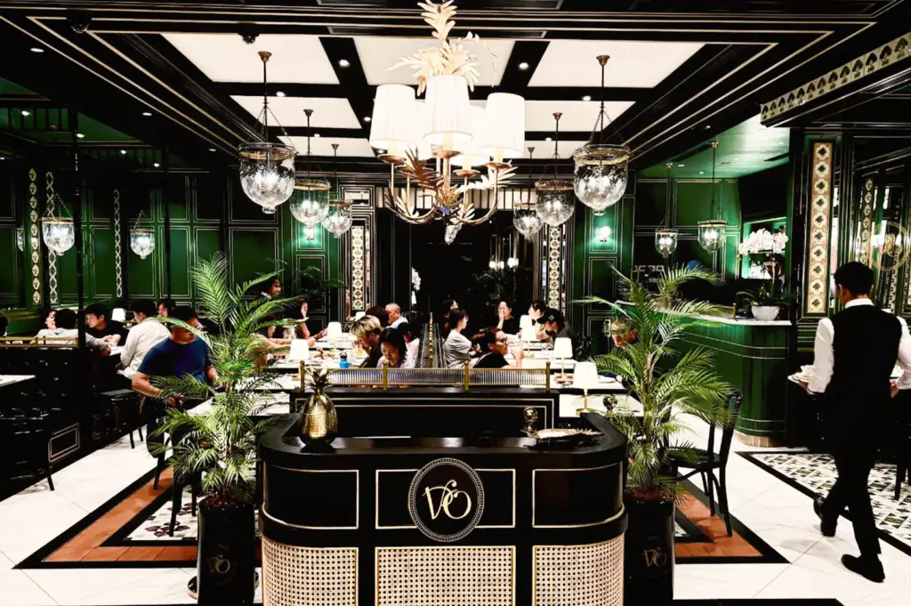 An elegant, brightly lit restaurant interior features patrons dining at tables flanked by vibrant emerald green walls with classic molding and two large potted palm plants. In the foreground, a dark wooden host stand prominently displays a gold circular "VO" logo and a golden pineapple decoration under an ornate, multi-bulb central chandelier and several hanging glass globes. On the right, a waiter dressed in a white shirt and black vest walks past the dining area, which is set against a black-and-white coffered ceiling and patterned tile flooring.