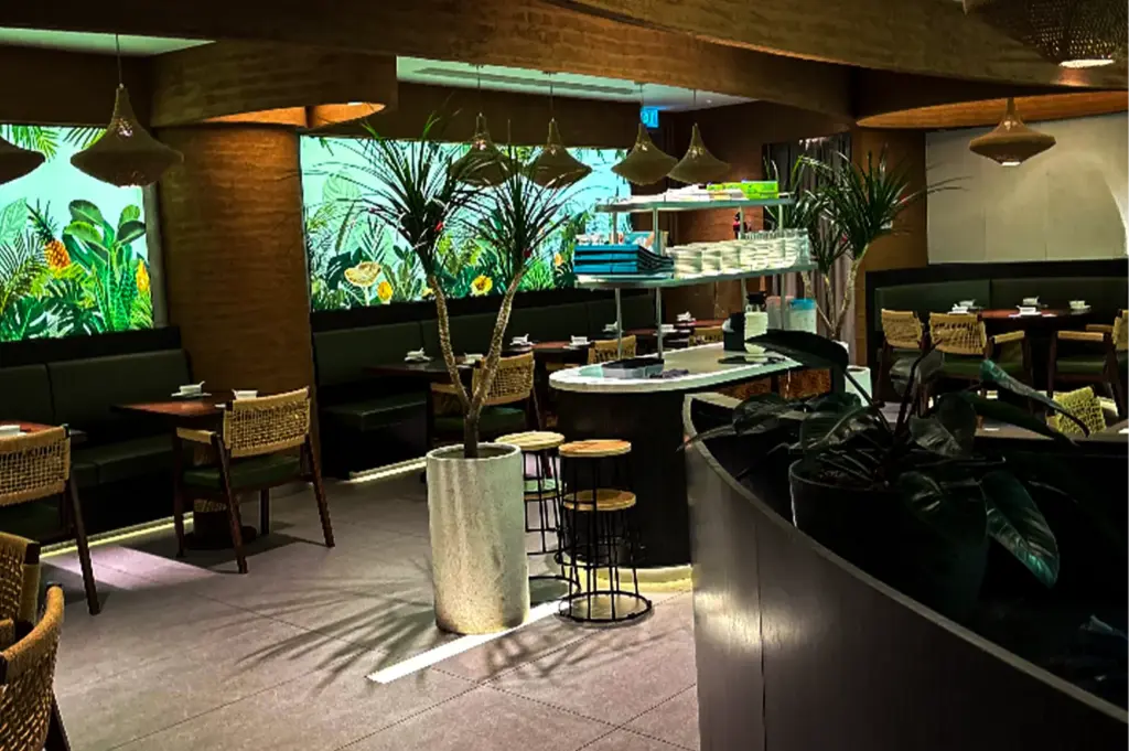 An interior view of a moody, tropically themed restaurant featuring warm wooden tables paired with woven wicker-back chairs and dark green banquette seating. The background highlights illuminated wall panels depicting a lush jungle of green leaves and yellow flowers, complemented by several potted indoor trees and plants throughout the space. Overhead, woven cone-shaped pendant lights cast a soft glow over the dining area and a central kitchen service station stocked with white plates.