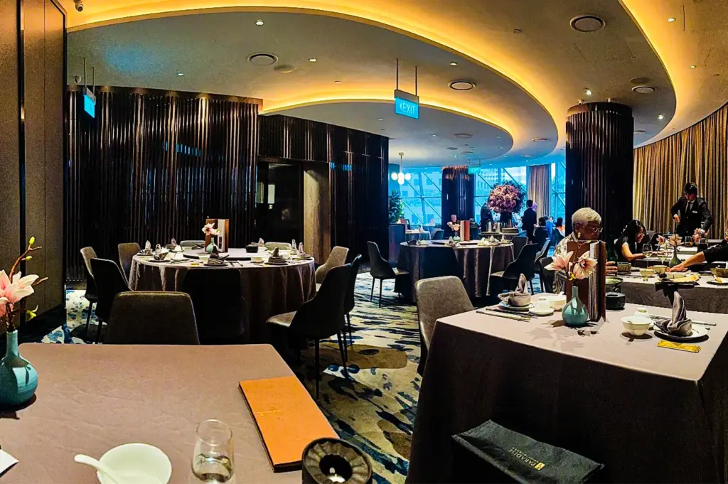 An interior view of a spacious, upscale restaurant featuring large round dining tables covered in grey-brown tablecloths and surrounded by dark modern armchairs. The room is designed with an elegant curved ceiling illuminated by warm recessed lighting, dark slatted wood accent walls, and a vibrant, patterned carpet in shades of blue, gold, and grey. In the background, patrons dine near large windows that offer a glimpse of the outdoors, while the foreground shows a detailed table setting with a small vase holding pink flowers, a menu booklet, and white tableware.