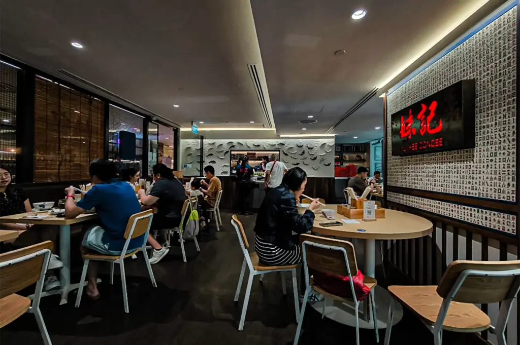 An interior view of a modern, casual restaurant called "MUI KEE CONGEE," where patrons sit at round and square light wood tables eating with chopsticks on a dark floor. The focal point on the right is a prominent wall section covered in a dense pattern of tiny, colorful icons resembling mahjong tiles, featuring a black rectangular sign with glowing red Chinese characters and the English name underneath. In the background, staff stand near a food prep counter flanked by a textured white wall decorated with a school of sculptural fish, while the left side features dark windows with roll-up bamboo blinds.