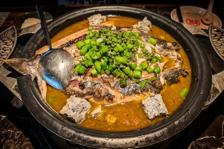 An overhead, close-up shot captures a whole grilled fish submerged in a bubbling, yellowish-green broth inside a large, heavy black metal pan, representing the image "1-Somerset Food The Ultimate Guide.webp". The center of the fish is piled high with a generous mound of chopped fresh green chilies and clusters of whole green peppercorns, while chunks of grayish tofu or spice paste are scattered along the edges of the pan. A large metal serving spoon rests on the left side of the dish, ready to serve the fiery, aromatic Sichuan-style meal.