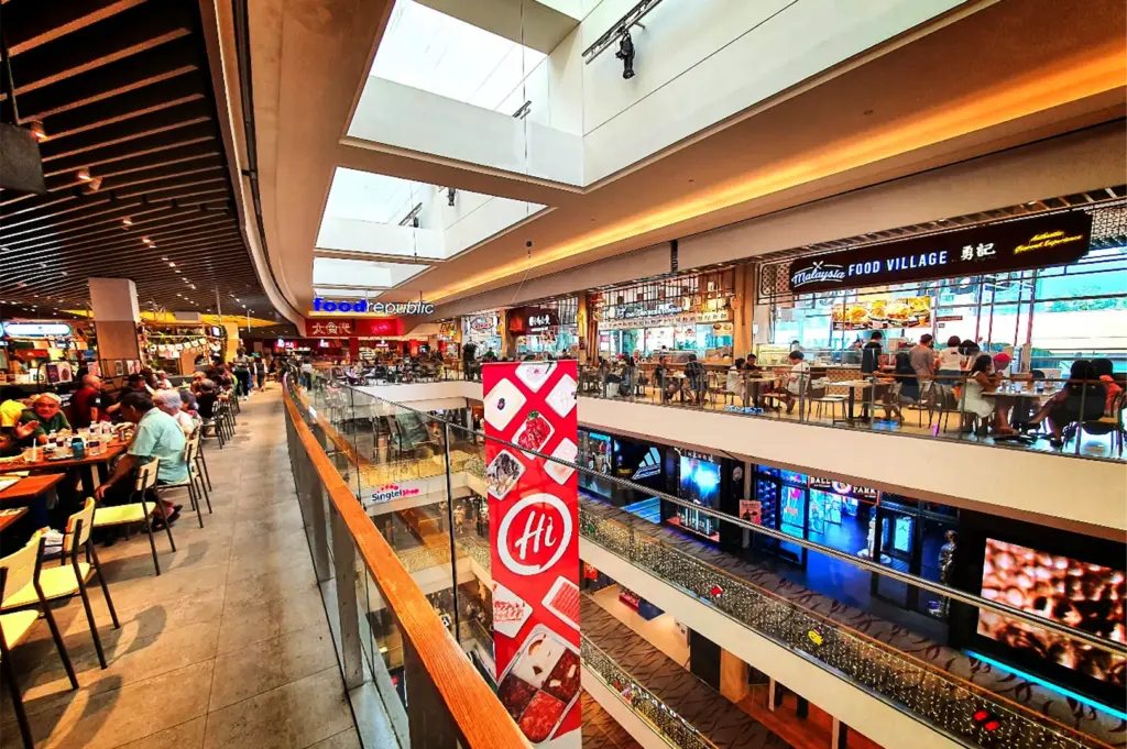 A high-angle, wide shot captures the bustling interior of a multi-level shopping mall's food court area, representing the image "5-Somerset Food The Ultimate Guide.webp". From an upper-level walkway lined with patrons dining at tables on the left, the view opens up to a multi-story atrium illuminated by large rectangular skylights above. Across the glass-railed balcony, various crowded eateries are visible, including a prominent "Food Republic" entrance in the distance and a "Malaysia Food Village" storefront to the right. A tall, red promotional banner with a circular "Hi" logo hangs centrally in the atrium, while retail shops on the lower floors can be seen down below.