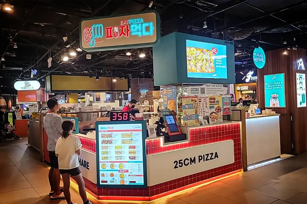 An eye-level, medium shot captures a modern food kiosk called "Eat Pizza" inside a bustling indoor mall basement, representing the image "6-Somerset Food The Ultimate Guide.webp". The kiosk features a prominent neon sign with both English and Korean text, a digital display screen showcasing a "Crispy Potato Bacon" pizza, and a base clad in glossy red square tiles with the text "25CM PIZZA" emblazoned on the front. A large, illuminated menu board stands at the corner, where a man in a striped shirt and a young girl are waiting to order, while a staff member in a face mask works behind the counter near neighboring stalls like "The Açai Affair."