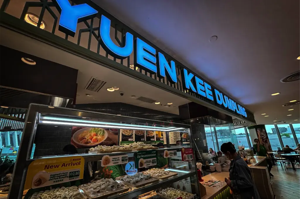 A low-angle, medium-wide shot captures the storefront of a restaurant named "YUEN KEE DUMPLING," represented in the image "7-Somerset Food The Ultimate Guide.webp". The shop features large, modern, blue-lit neon block letters spelling out its name above a glass-enclosed preparation counter and display case. Inside the glass display, numerous trays filled with raw, neatly arranged dumplings are showcased alongside promotional signs like "New Arrival" and "Our Recommendation." A woman stands at the counter on the right ready to order, while the restaurant's indoor dining area extends into the background, brightened by natural light from windows looking out onto an urban backdrop.