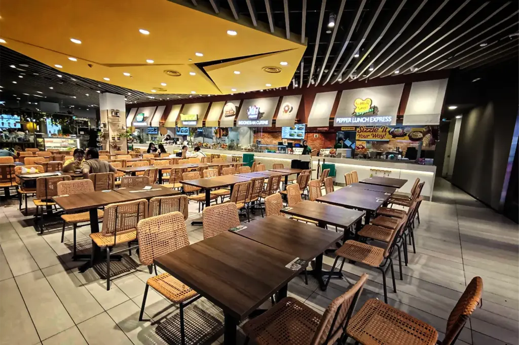 A wide, eye-level shot captures a bright and spacious indoor food court dining area, represented in the image "8-Somerset Food The Ultimate Guide.webp". The foreground features neatly aligned rows of dark wooden tables paired with light brown woven wicker chairs, with only a few patrons scattered toward the back. The ceiling is modernly designed with white acoustic slats and a prominent geometric, yellow-lit dropped ceiling section on the left. In the background, a row of food stalls with white awnings includes visible signs for "Indonesian Cuisine" and "Pepper Lunch Express," showcasing a clean, welcoming environment.