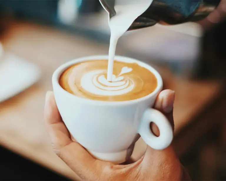 Fresh milk being carefully poured into a cup of espresso, creating delicate latte art patterns on the creamy surface. The swirl of milk blends smoothly with the rich, dark coffee, highlighting the craft and precision of the barista.