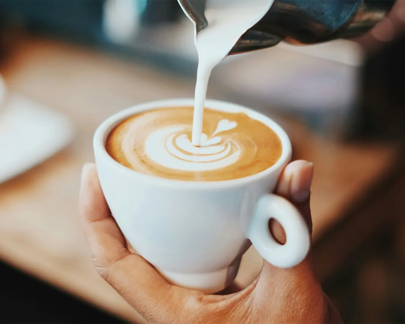 Fresh milk being carefully poured into a cup of espresso, creating delicate latte art patterns on the creamy surface. The swirl of milk blends smoothly with the rich, dark coffee, highlighting the craft and precision of the barista.