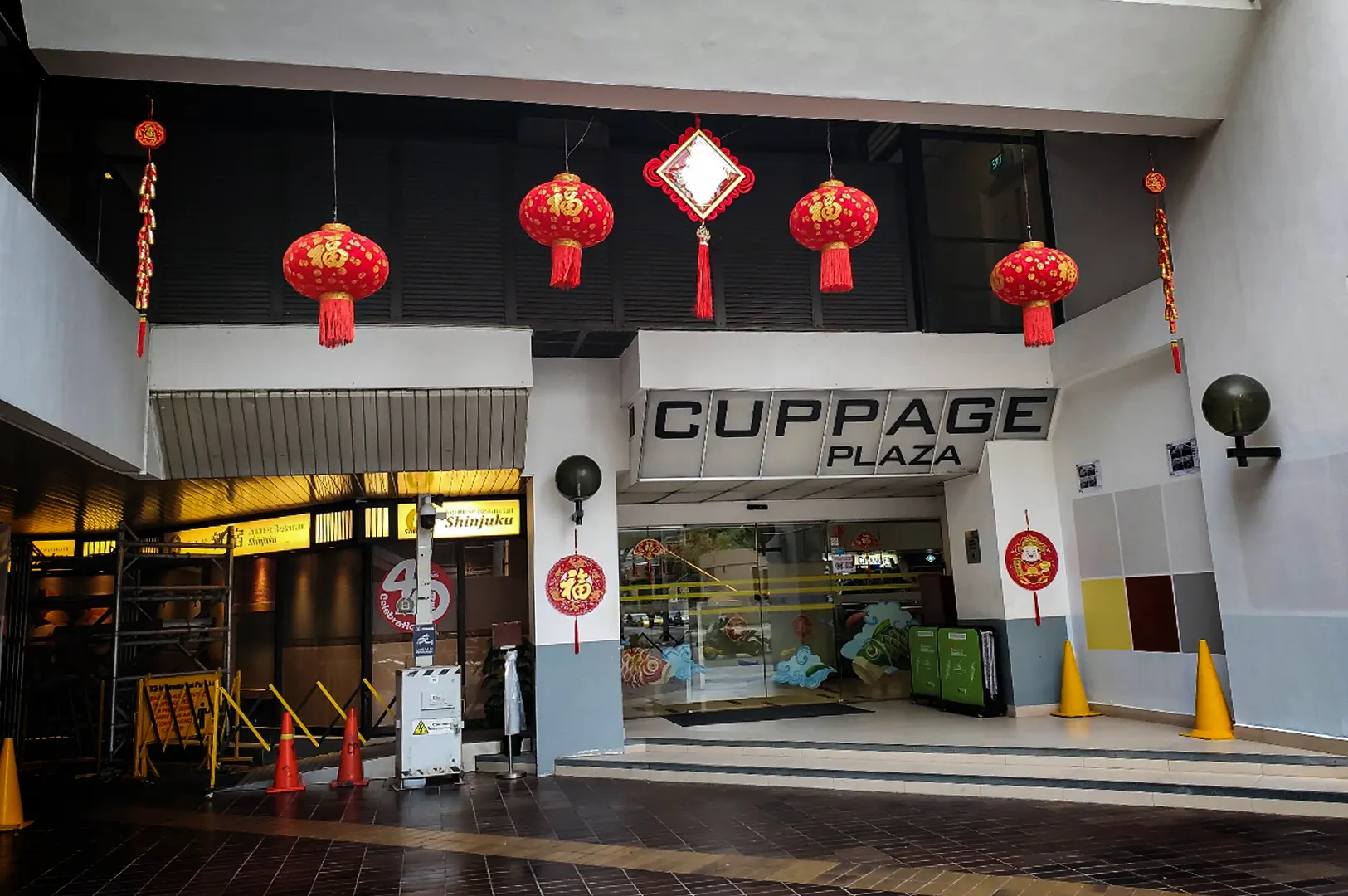 An eye-level, medium shot captures the concrete entrance to the "CUPPAGE PLAZA" commercial building, representing the file "1-Discover Cuppage Plaza Food.webp". The building features prominent block letters spelling out its name above a set of glass sliding doors, which are decorated with colorful ocean wave and fish illustrations. Festooned with Chinese New Year decorations, several large red paper lanterns with gold calligraphy and tassels hang from the overhead overhang, complemented by circular red cutout banners on the adjacent pillars. To the left, a dimly lit driveway or walkway leads past a yellow sign for a restaurant named "Shinjuku," while orange and yellow traffic cones line the tiled ground.