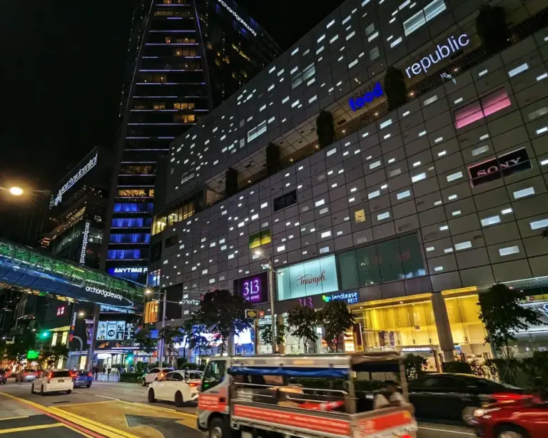 Exterior view of 313@Somerset shopping mall along Orchard Road in Singapore at night, brightly lit with modern glass architecture, glowing storefronts, and city lights reflecting the vibrant nightlife of the Orchard shopping district.