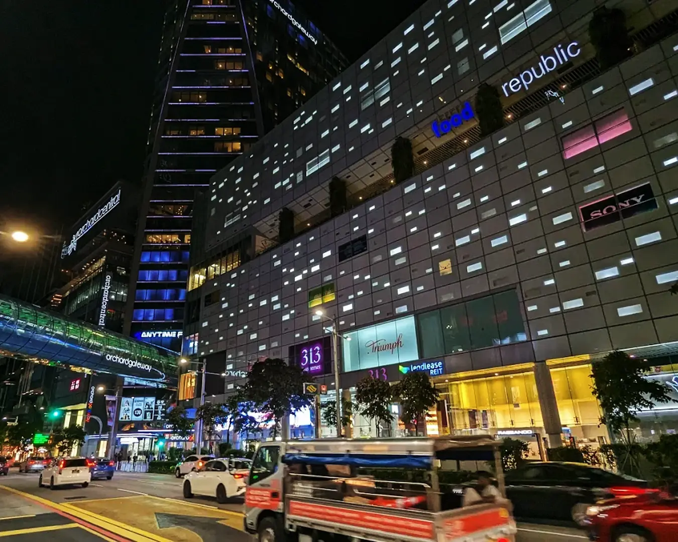 Exterior view of 313@Somerset shopping mall along Orchard Road in Singapore at night, brightly lit with modern glass architecture, glowing storefronts, and city lights reflecting the vibrant nightlife of the Orchard shopping district.