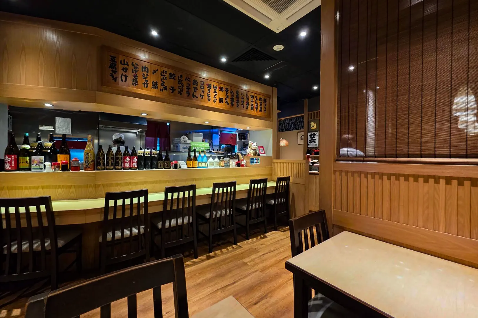 The warm, wooden interior of a traditional Japanese restaurant features a counter lined with dark wooden chairs and an array of sake bottles. Traditional wooden menu plaques hang above the kitchen counter, complementing the light wood paneling and warm ambient lighting throughout the dining area.
