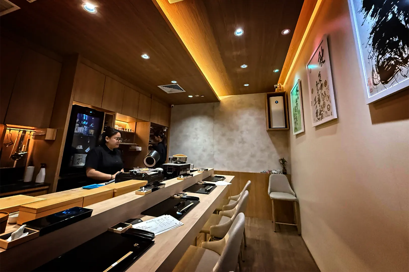 An eye-level, wide shot captures the intimate interior of a cozy, narrow Japanese restaurant, representing the file "2-Discover Cuppage Plaza Food.webp". The space is defined by a long, light wooden counter bar paired with several upholstered cream chairs, each station neatly preset with black placemats and chopsticks. Behind the counter, a female staff member in a black uniform stands ready near kitchen equipment and wooden cabinetry, while another employee is visible in the background. The warm ambiance is enhanced by a wood-paneled ceiling with recessed cove lighting, textured beige walls adorned with framed Japanese calligraphy, and a solitary accent chair tucked in the far corner.
