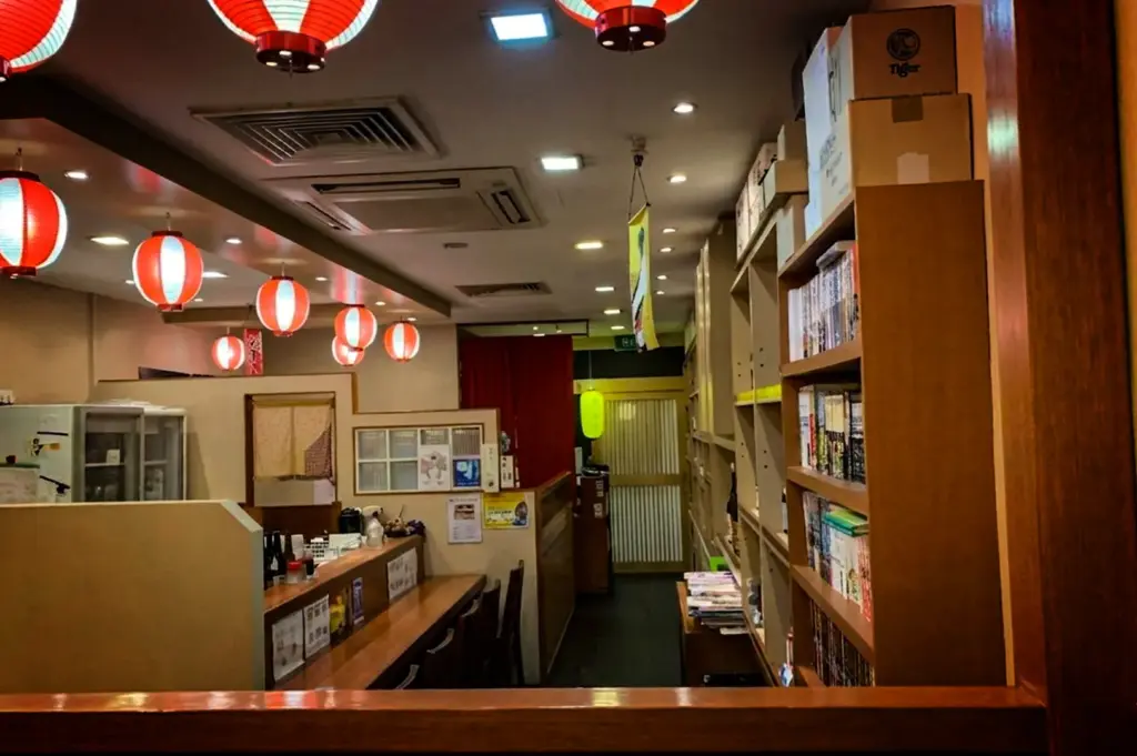 An eye-level, medium shot captures the warm and slightly cluttered interior of an authentic Japanese restaurant, representing the file "3-Discover Cuppage Plaza Food.webp". A row of glowing red-and-white paper lanterns hangs along the ceiling, illuminating a wooden counter bar with a few low stools on the left. To the right, a tall wooden bookshelf is filled with numerous volumes of manga or books, topped with cardboard shipping boxes. In the background, a narrow hallway is framed by low partitions, a red fabric curtain, and a traditional sliding door, creating a cozy, lived-in Izakaya atmosphere.