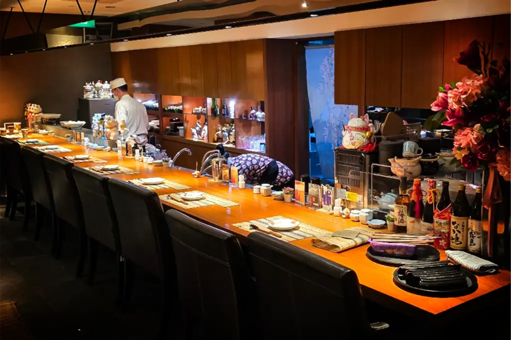 An eye-level, medium shot captures the warm, inviting interior of a high-end Japanese dining bar, representing the file "4-Discover Cuppage Plaza Food.webp". A long, polished wooden counter stretches diagonally across the frame, lined with sleek black leather dining chairs and meticulous individual place settings with woven placemats. Behind the counter, a chef in a white uniform works in the back, while a server in a traditional patterned kimono leans over to prepare a station. The background features rich wooden cabinetry displaying assorted bottles and a lucky cat figure, balanced on the right by a large, vibrant bouquet of pink and red flowers next to a selection of premium sake bottles.