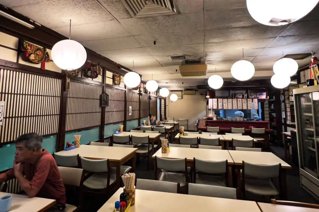 An eye-level, wide shot captures the traditional interior of an Izakaya-style Japanese restaurant, representing the file "5-Discover Cuppage Plaza Food.webp". The dining area is filled with neat rows of wooden tables paired with light gray upholstered chairs, each set with containers of wooden chopsticks and condiments. Overhead, numerous large, glowing white paper lantern globes hang from a textured acoustic tile ceiling, casting a warm light throughout the space. The left wall is decorated with classic wooden latticework shoji panels over a teal accent base, while an older man in a reddish-brown shirt sits in the lower-left foreground looking toward a small open kitchen counter in the background, where traditional menu banners hang.