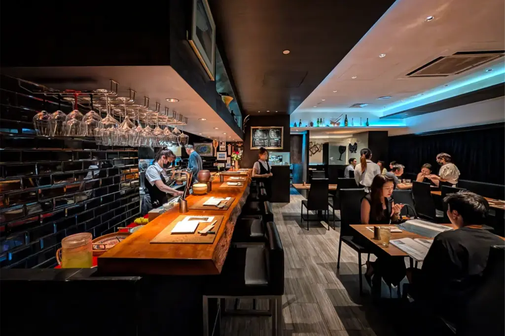 An eye-level, wide shot captures the sleek and modern interior of a dimly lit, upscale Japanese restaurant, representing the file "6-Discover Cuppage Plaza Food.webp". On the left, a long, thick live-edge wooden counter bar spans the depth of the room, illuminated by overhead spotlights and set with low stools, while a staff member in a face mask works behind it beneath rows of upside-down hanging wine glasses. To the right, several small wooden tables are occupied by patrons dining in high-backed black chairs. The contemporary space features dark wood-plank flooring, a textured black tiled accent wall behind the bar, and a recessed ceiling with cool-toned blue cove lighting that adds a sophisticated mood to the dining area.