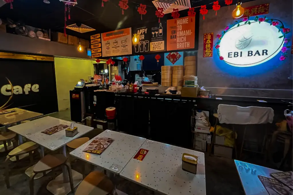 An eye-level, medium-wide shot captures the eclectic, dimly lit interior of a restaurant named "EBI BAR," representing the file "7-Discover Cuppage Plaza Food.webp". In the foreground, white speckled tables paired with simple wooden stools sit empty, each set with menus and tissue boxes. In the background, a dark service counter is decorated with stacks of bamboo dim sum steamers, small hanging red paper lanterns, and Chinese New Year "Fu" character banners. Above the counter, colorful graphic signs advertise "Happy Hour" and "Umami-Rich Broth," while a large, oval, backlit sign with a blue glow prominently displays the restaurant's logo on a textured concrete wall to the right.