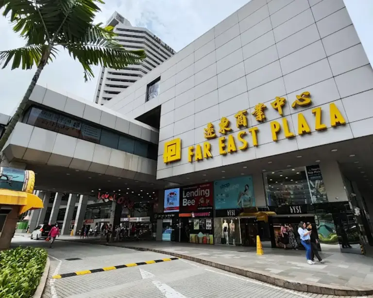 A bustling entrance to Far East Plaza features a large yellow sign and multiple shops. A palm tree adds a tropical touch, under a partly cloudy sky.