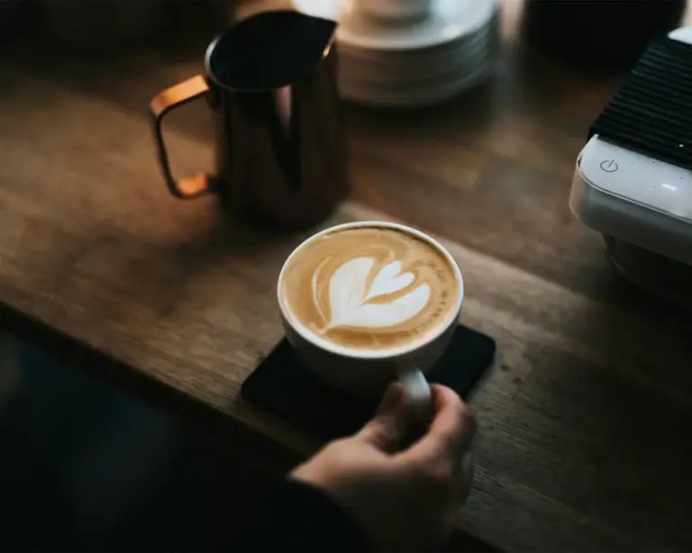 A hand holds a white cup of latte with intricate heart-shaped latte art on a wooden table. A metal pitcher and espresso machine are in the background, exuding a cozy café vibe.
