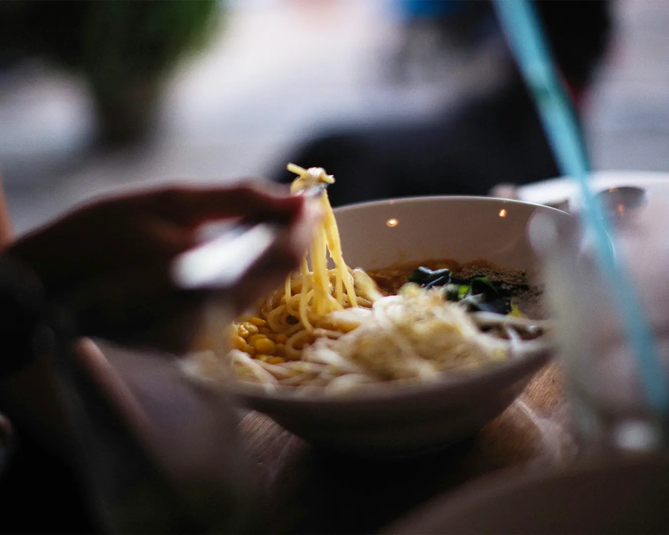 A person uses chopsticks to lift noodles from a bowl of ramen, with blurred background. Warm and cozy ambiance, suggesting a casual dining setting.