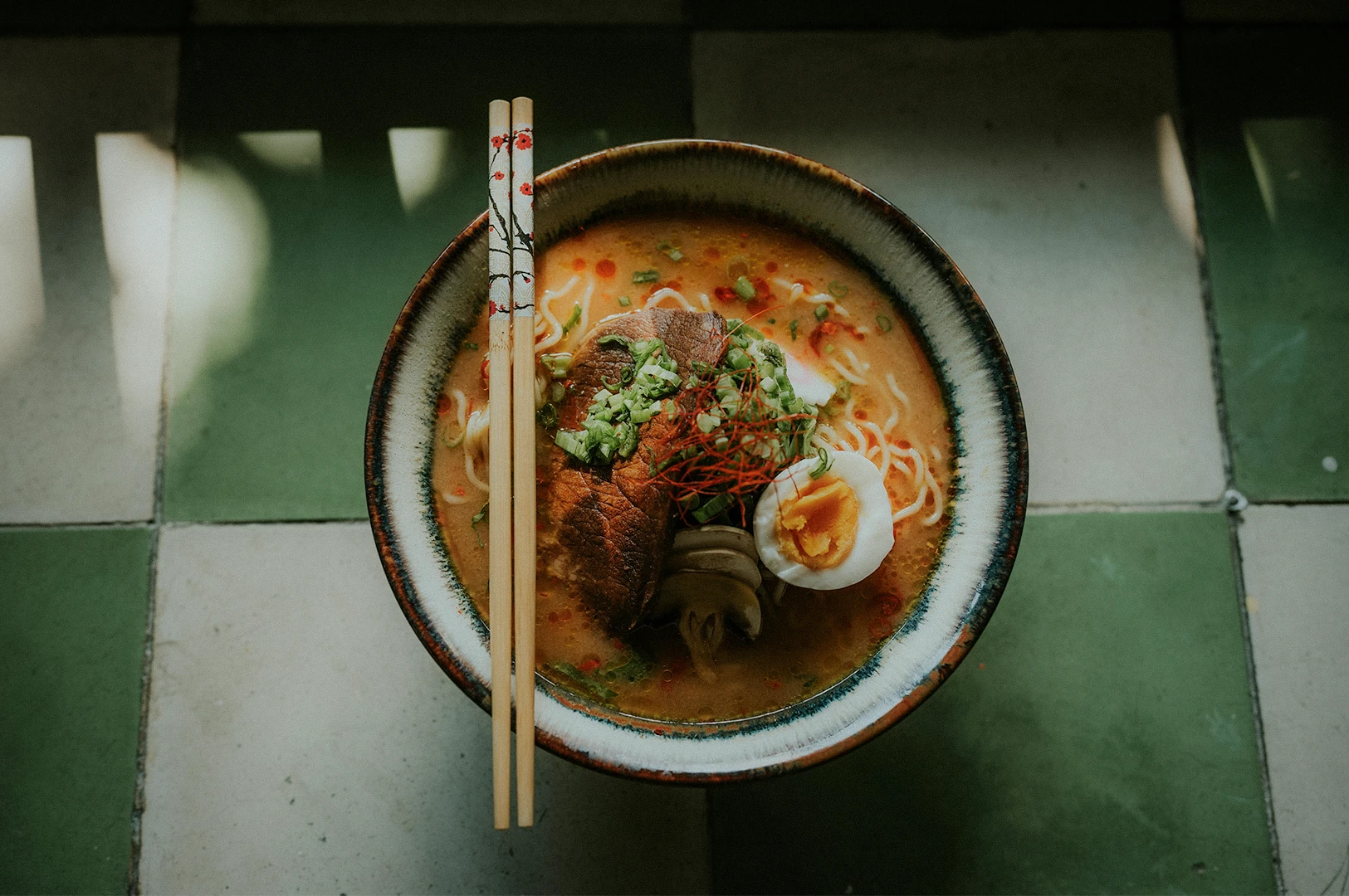 Authentic bowl of Japanese ramen with rich broth, tender chashu pork, a soft-boiled ajitama egg, and green onions, served with floral-patterned chopsticks on a vintage checkered tile floor.