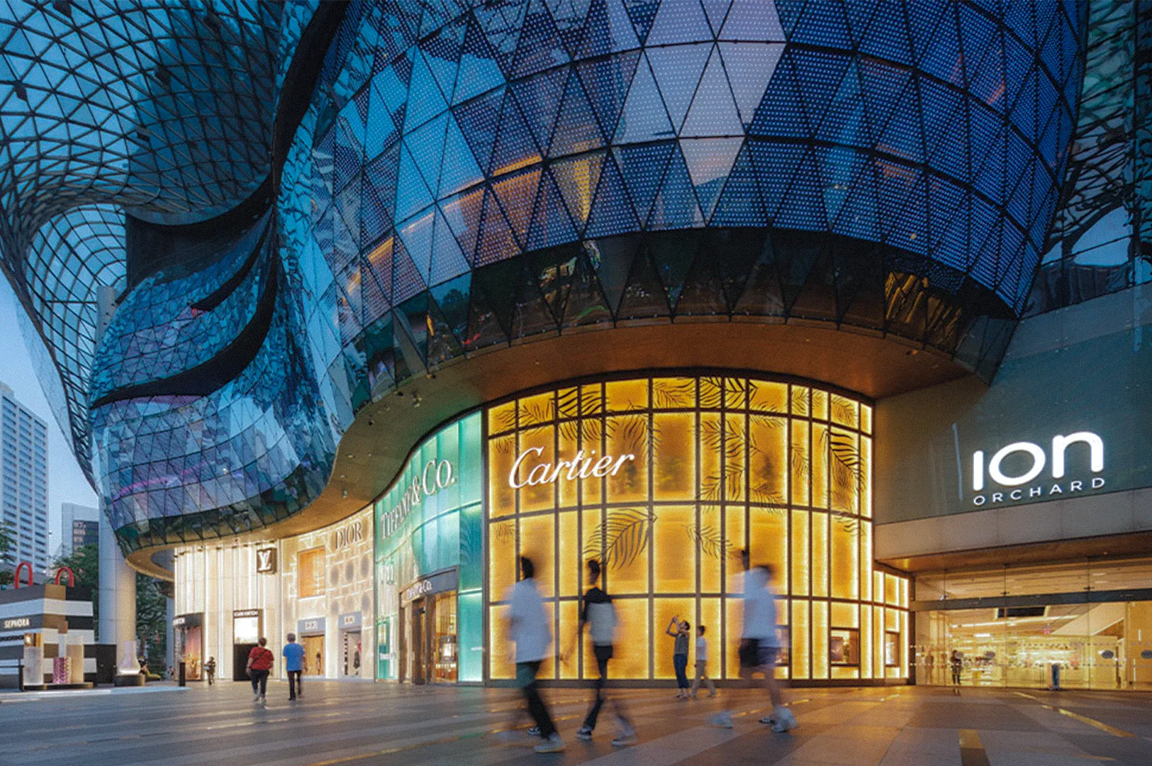An illuminated view of the ION Orchard luxury shopping mall in Singapore at dusk, featuring high-end retail storefronts for Cartier, Tiffany & Co., Dior, and Louis Vuitton under a modern glass architecture.