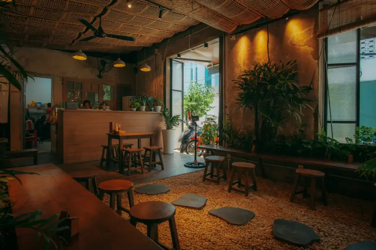 Chic cafe interior featuring a unique pebble floor with stone pavers, woven rattan ceiling panels, warm lighting and rusing wooden furniture surrounded by lush indoor plants for touch of tropical vibe.