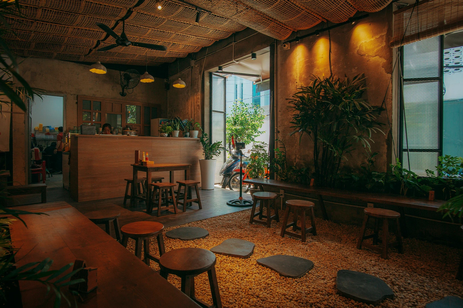 Chic cafe interior featuring a unique pebble floor with stone pavers, woven rattan ceiling panels, warm lighting and rusing wooden furniture surrounded by lush indoor plants for touch of tropical vibe.