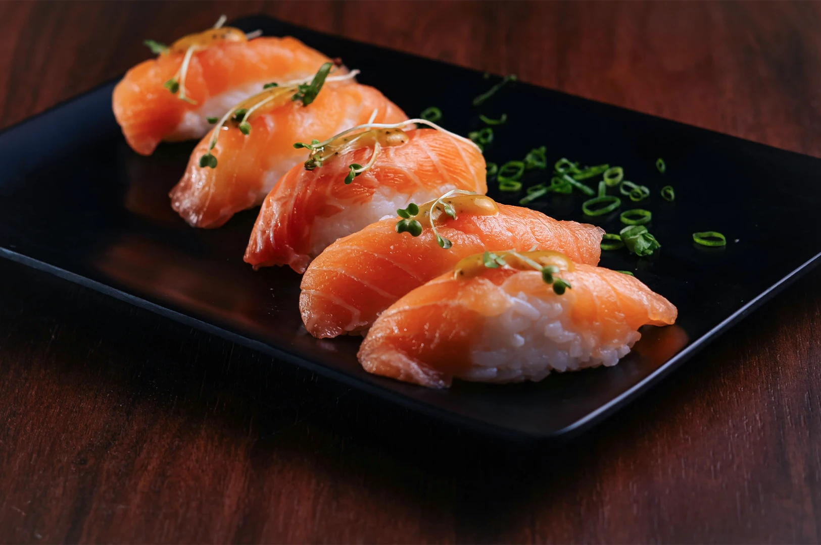 Five pieces of fresh salmon nigiri sushi elegantly plated on a black ceramic dish with microgreens and garnishes. Gourmet Japanese cuisine photography for restaurant menus and food blogs.