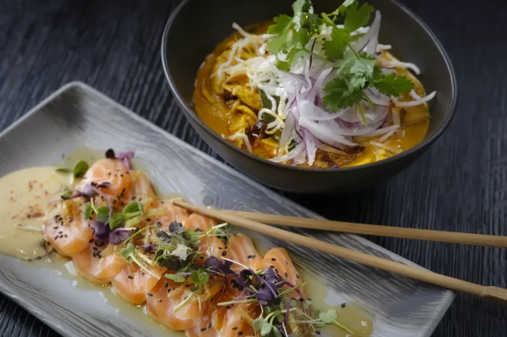 Elegant Japanese food spread featuring fresh salmon carpaccio with microgreens and a bowl of spicy ramen topped with herbs. Gourmet fusion cuisine photography.