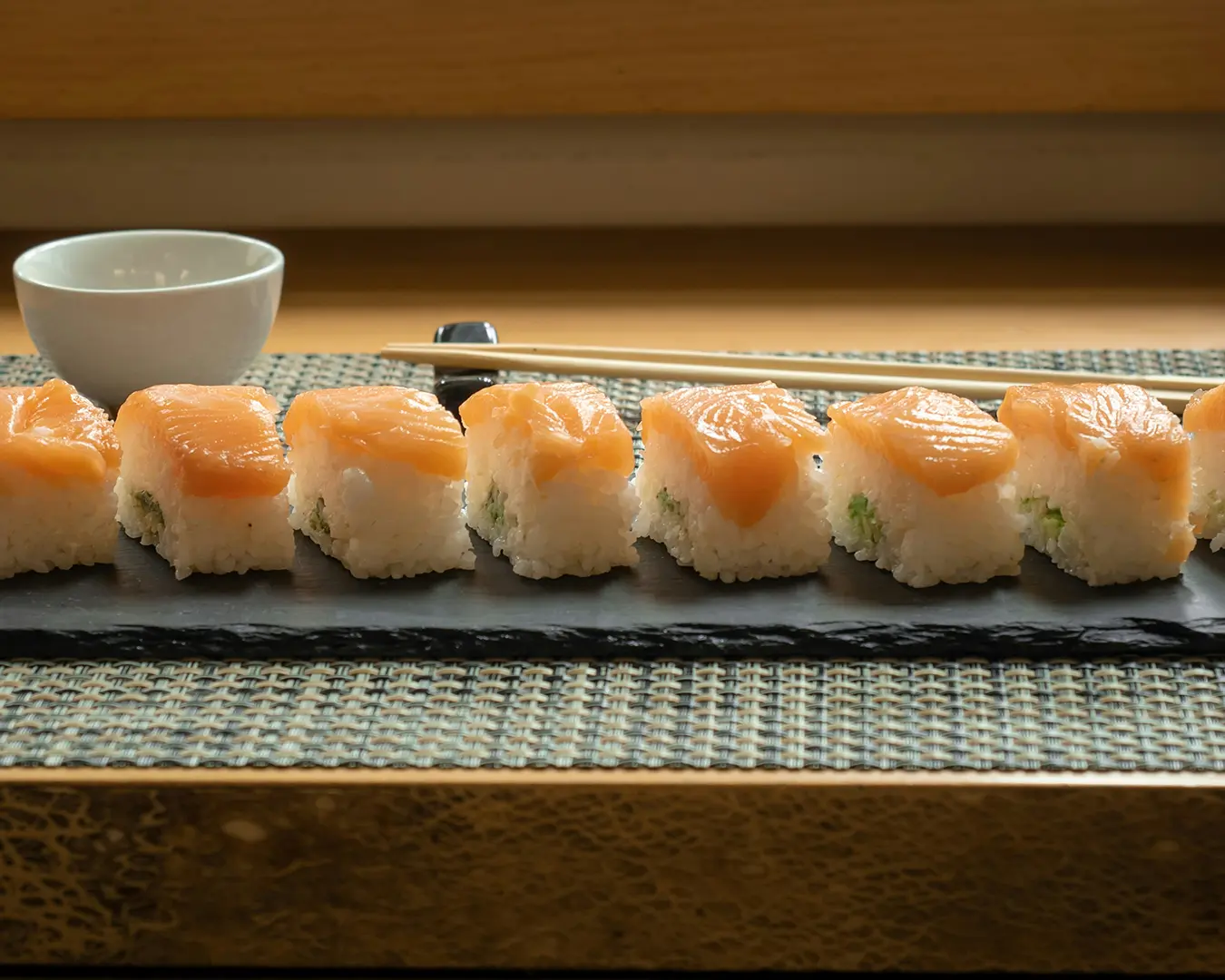 Sushi pieces topped with salmon are neatly arranged on a black slate. Nearby, there’s a dish of soy sauce, pickled ginger, and a pair of chopsticks, creating an inviting and elegant dining setting.