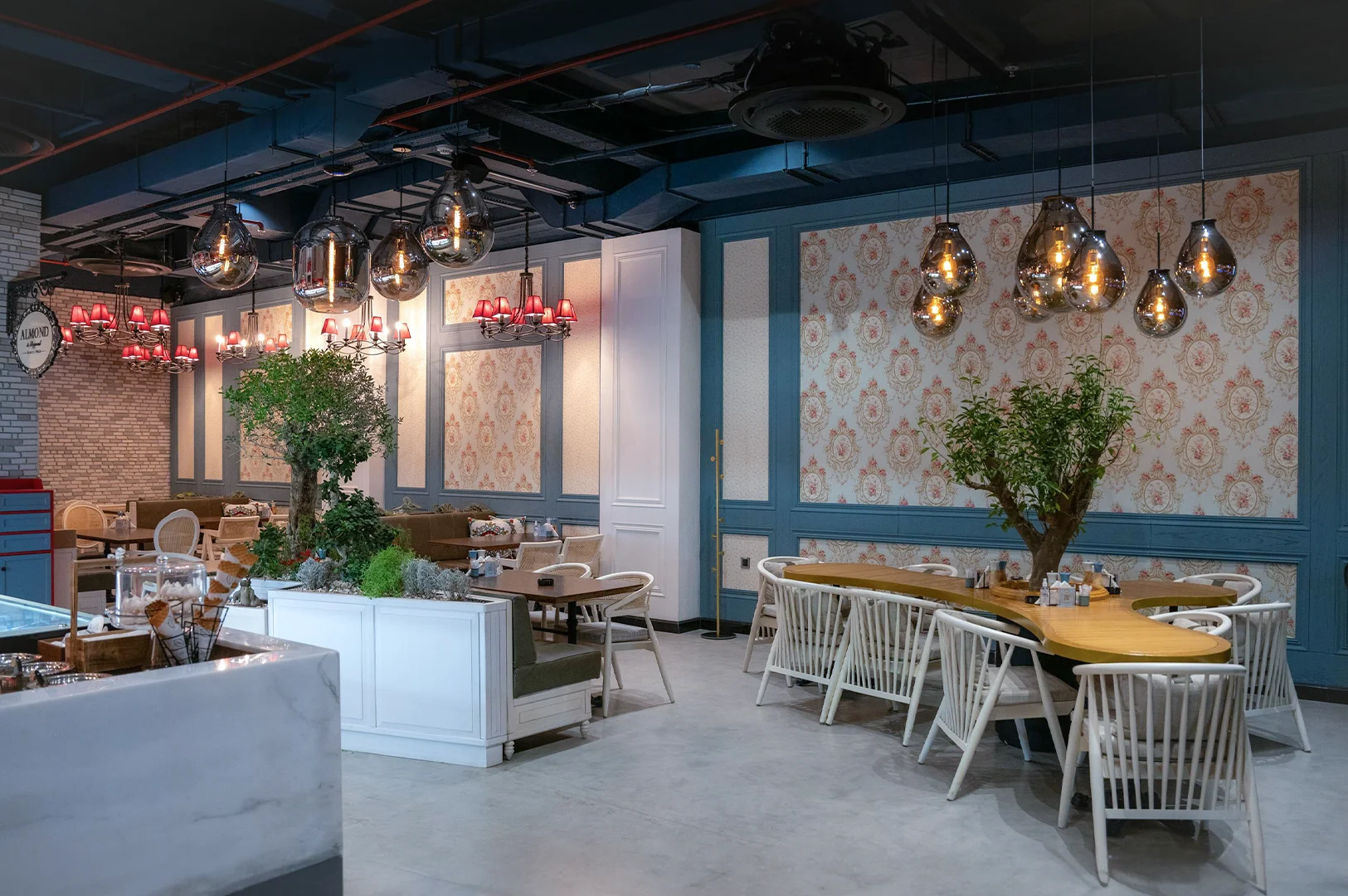 Luxurious cafe interior with floral wallpaper, blue wainscoting, curved wooden table, pendant lights, and greenery ambiance