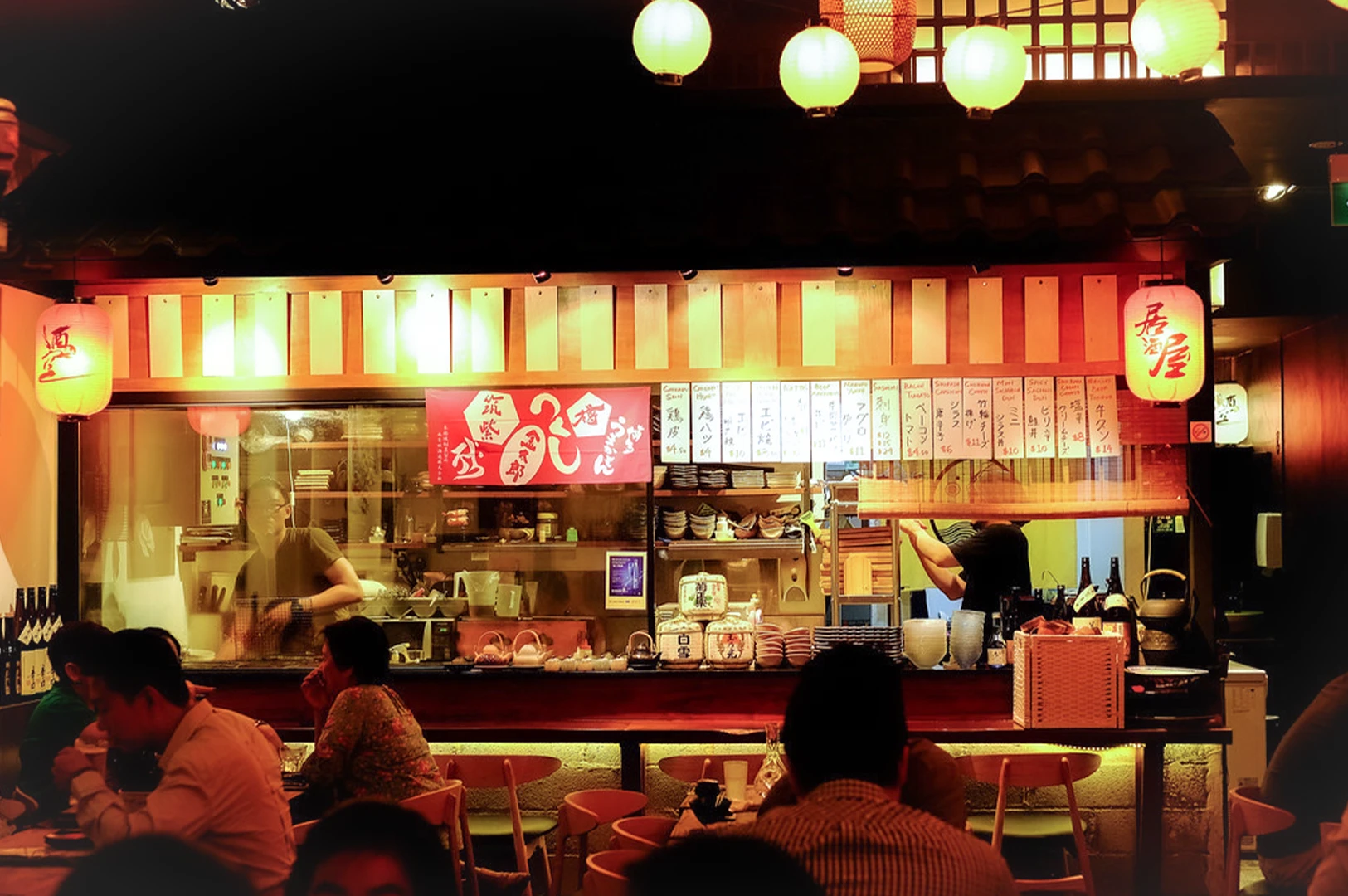 Cozy interior of Shukuu Japanese Izakaya and Bar in Singapore featuring glowing paper lanterns, a busy open kitchen, and diners. Authentic Japanese dining atmosphere on Stanley Street for SEO.