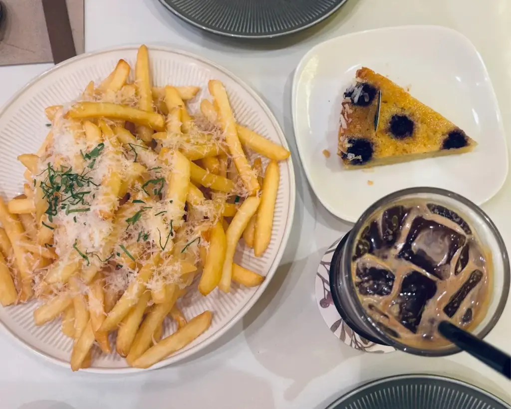 A plate of golden fries topped with cheese and herbs, a slice of blueberry cake on a square plate, and an iced coffee with a straw on a floral coaster.