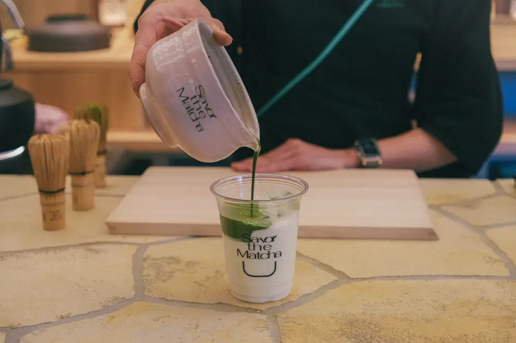 A barista pouring a vibrant green concentrated matcha tea from a ceramic bowl into a plastic cup filled with iced milk, creating a layered matcha latte at a cafe.