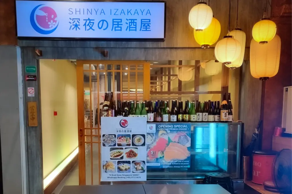 Restaurant entrance for Shinya Izakaya at 79 Boat Quay, Singapore, featuring traditional paper lanterns, a collection of Japanese sake bottles, promotional posters, and Japanese menu text.