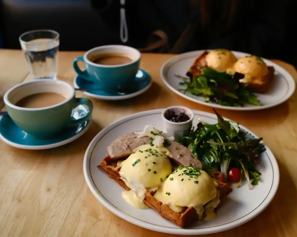 Plates of eggs Benedict with waffles, sausage, and greens on a wooden table. Teal cups of coffee and water glass add a cozy brunch ambiance.