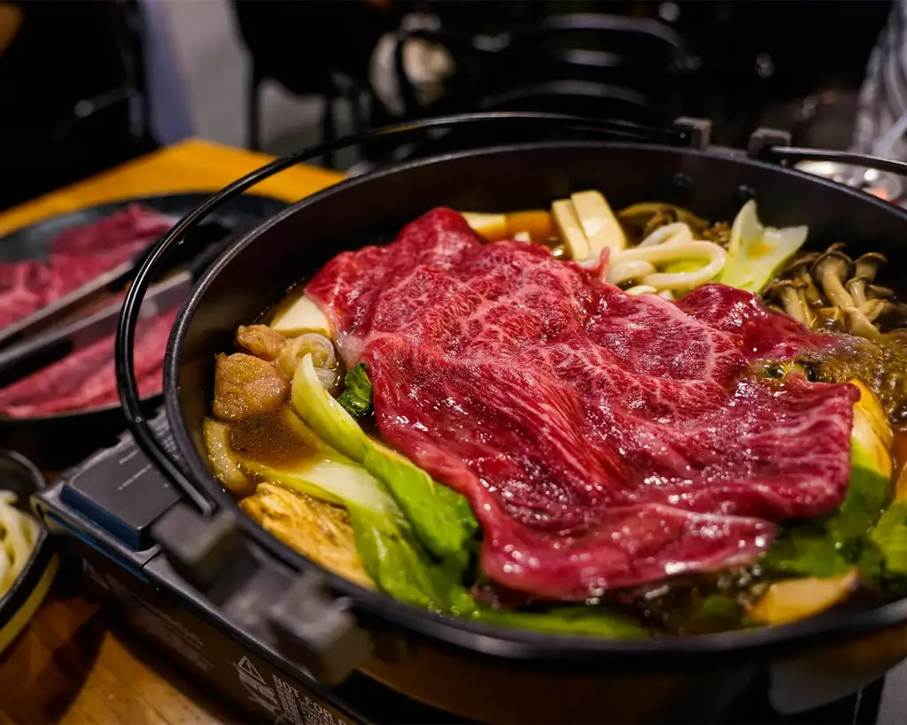 Close-up of a hot pot with marbled beef slices, mushrooms, tofu, and cabbage simmering in broth. The scene conveys warmth and indulgence.