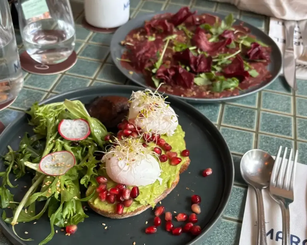 Poached eggs on avocado toast with pomegranate seeds and greens, next to a carpaccio-style salad. Fresh and vibrant breakfast spread.