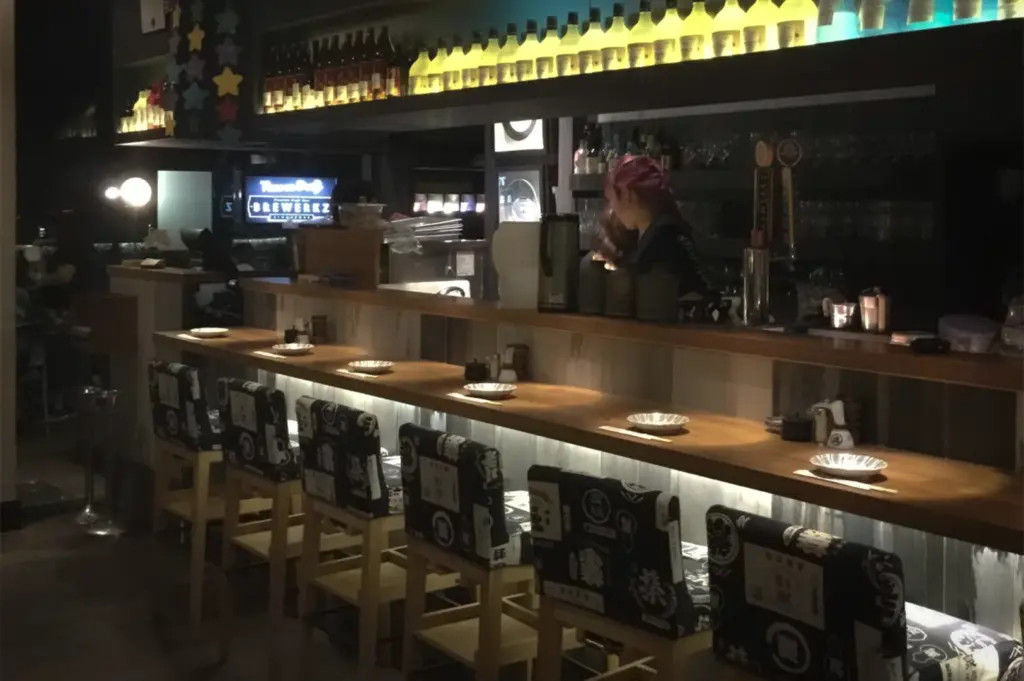 Interior bar counter of Shunjuu Izakaya at Robertson Quay, Singapore. Features wooden seating with patterned cushions, back-lit sake bottles, and authentic Japanese pub ambiance for local dining.