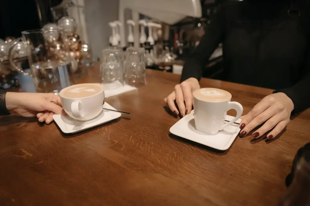 Two friends enjoying latte art in white ceramic cups at a wooden cafe bar. The cozy scene features a barista serving fresh coffee, perfect for cafe culture, morning routines, and social gatherings.