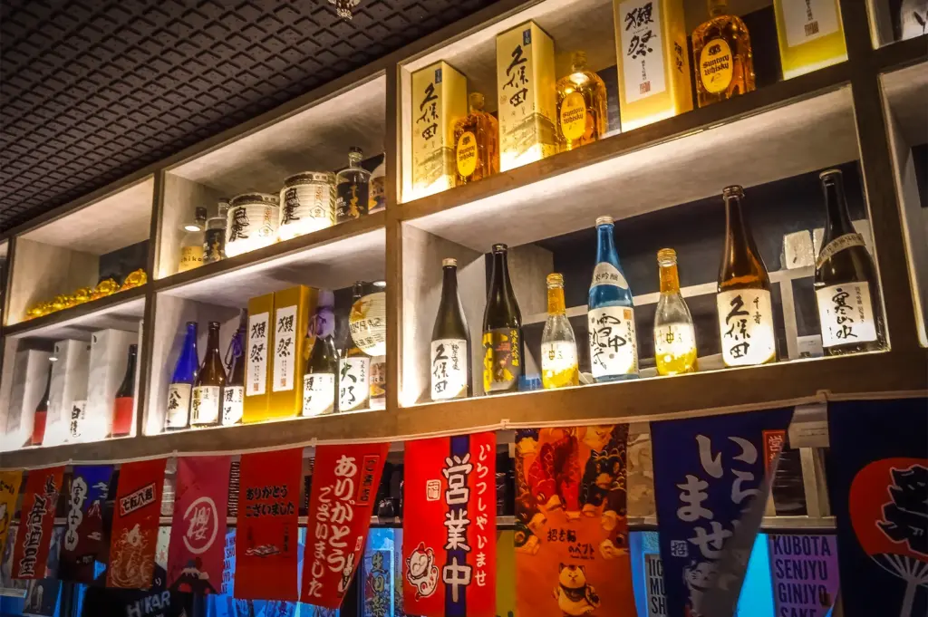 Display of premium Japanese sake and Suntory whisky on backlit shelves at Izakaya Hikari, Middle Road. Features traditional colorful noren banners and authentic decor for Singapore dining SEO.