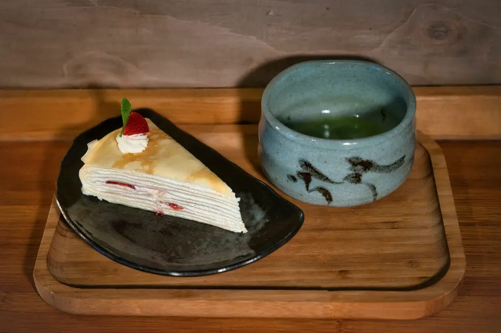 A slice of Japanese strawberry mille crepe cake on a black ceramic plate served with a bowl of hot matcha green tea on a wooden tray, featuring elegant cafe dessert presentation.