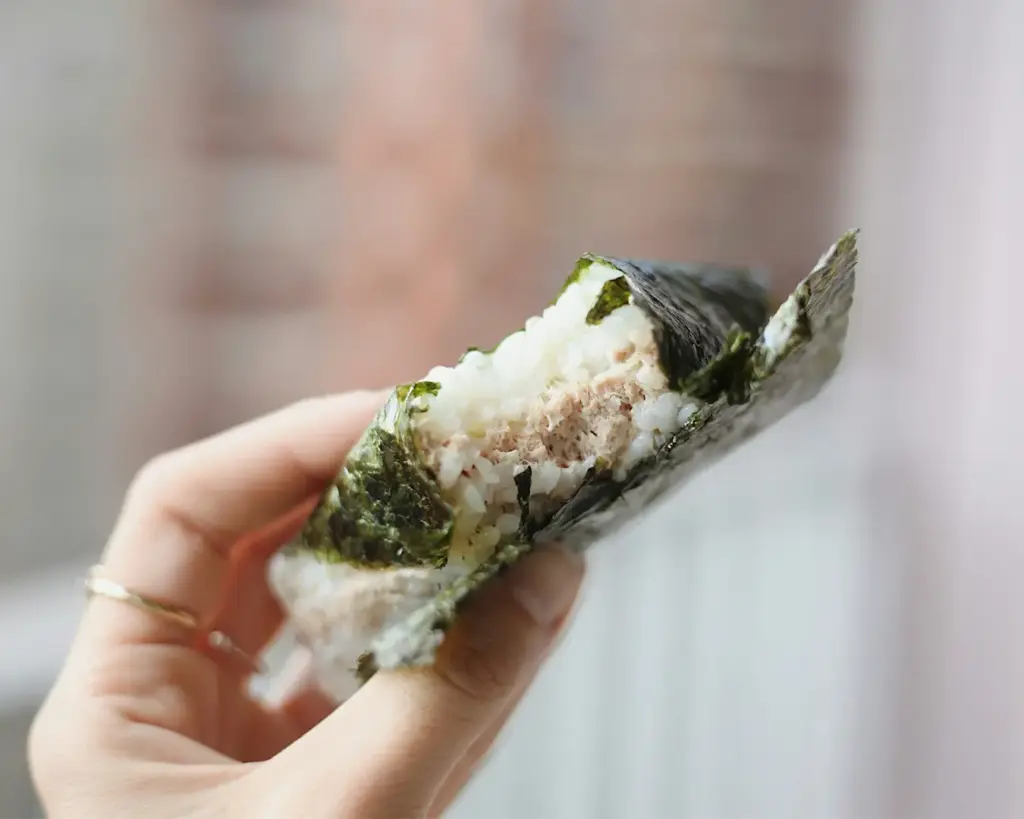 A hand holds a partially eaten onigiri, a Japanese rice wrapped in seaweed with a visible tuna filling, evoking a sense of freshness and simplicity.