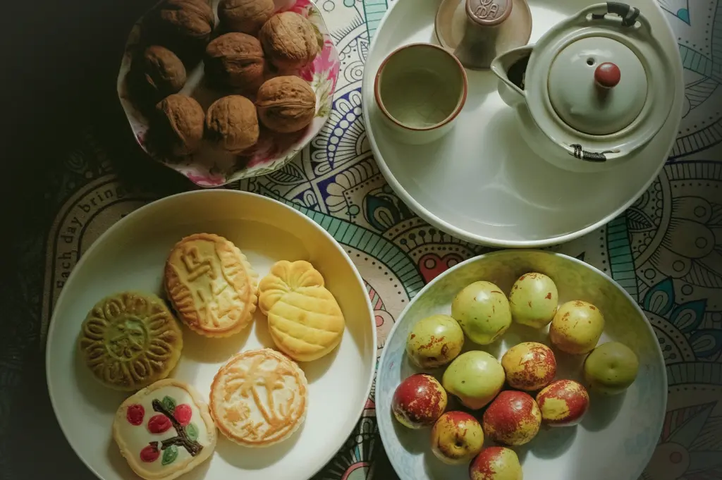 Assortment of Chinese mooncakes, fresh green and red jujube fruits, whole walnuts, and a traditional ceramic tea set on a patterned tablecloth, perfect for Mid-Autumn Festival celebrations.