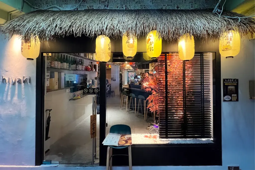 Exterior of Hiyaki Yakitori Izakaya Bar at Kampong Bahru featuring a thatched roof, glowing yellow lanterns, and a glimpse of the modern interior. Authentic Japanese grill and bar in Singapore